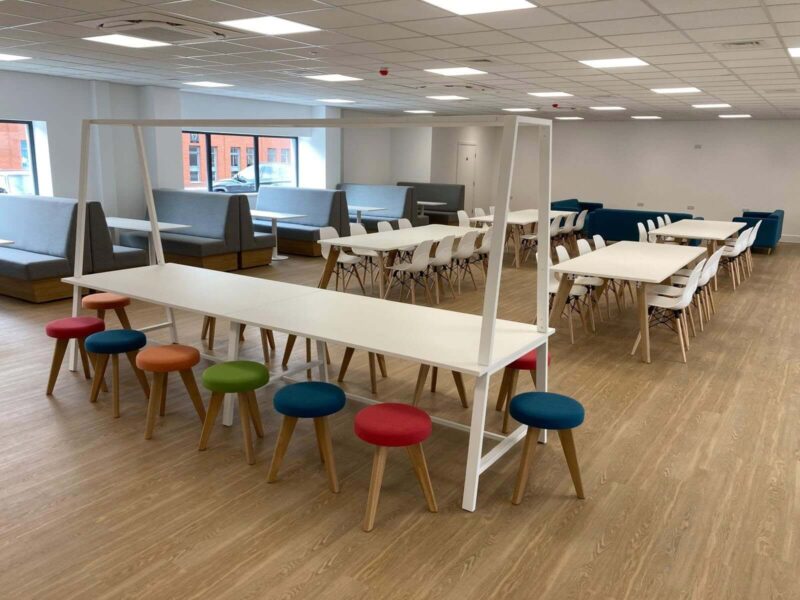 Wide view of an open-plan office breakout area featuring a white bench table with colourful Timba stools from Richard Butler Furniture in the foreground, rows of white dining tables with white chairs, and grey and blue booth seating along the walls.