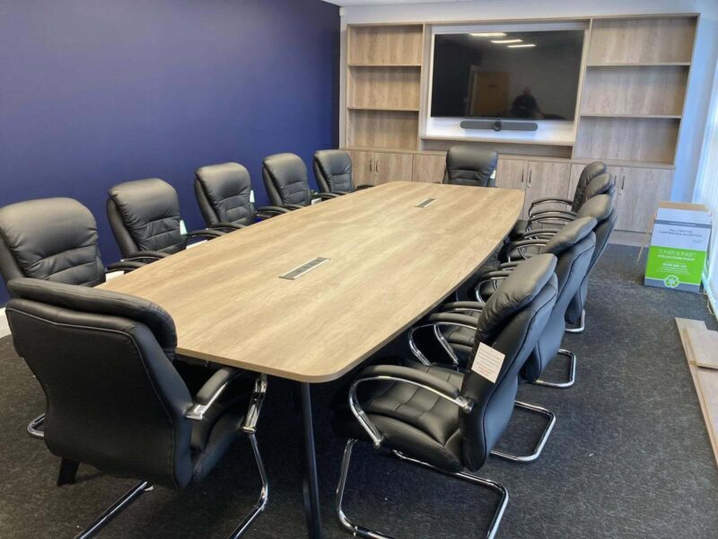 Executive boardroom with a large light oak boat-shaped conference table, black leather boardroom chairs with chrome frames, and a built-in oak media wall with wall-mounted TV screen and video conferencing camera, set against a navy blue feature wall.